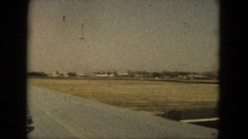 Airplane-window view at Acapulco airport in 1962, looking across runway pavement, open ground, and distant airport buildings.