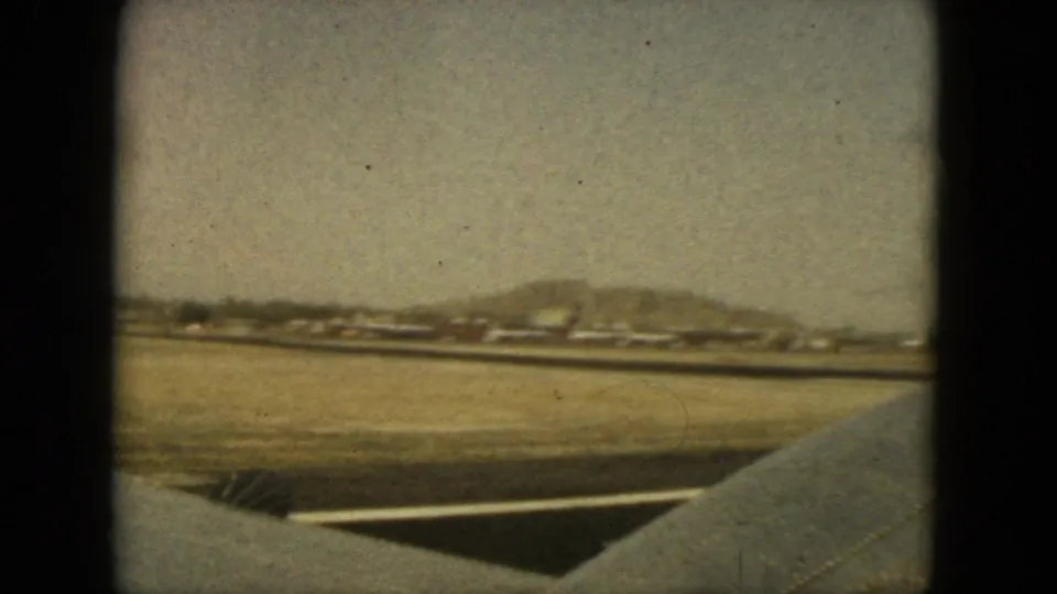 Airplane-window frame from 1962 Acapulco showing wing edge, runway markings, distant airport buildings, and low hills under haze.