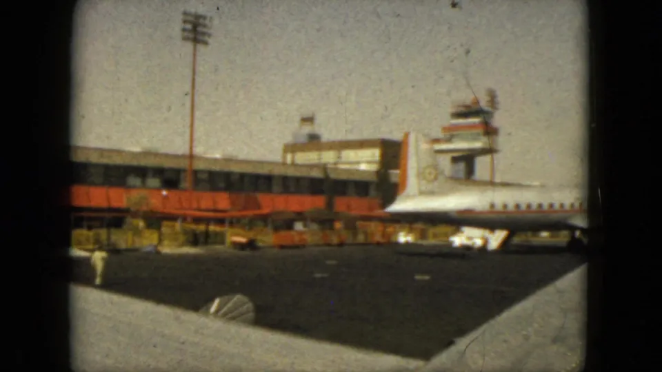 1962 Acapulco airport view with a white jet parked near the terminal, orange awnings, parked cars, and aircraft wing edge in the foreground.