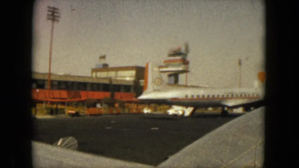 1962 Acapulco airport tarmac with a parked white jet, terminal building, orange awnings, ramp area, and wing edge in the foreground.