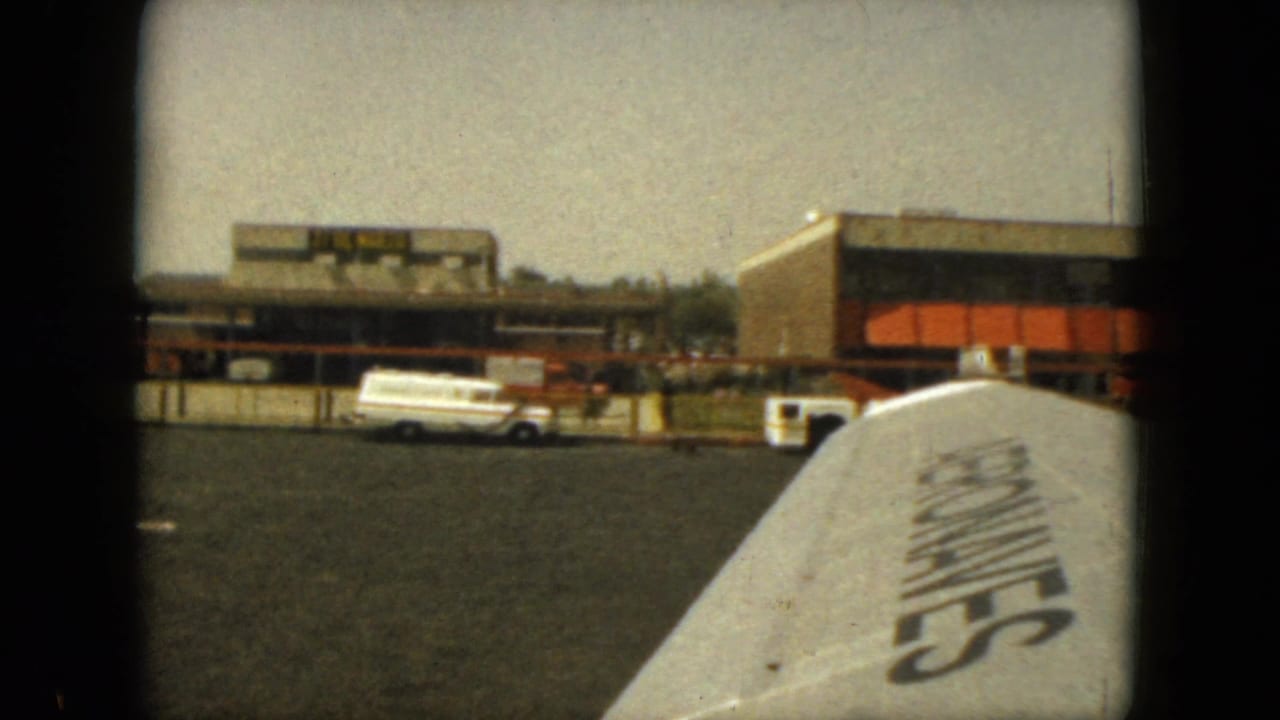 1962 Acapulco airplane-window view of a wing on the tarmac, with a low airport building and white service van in the background.