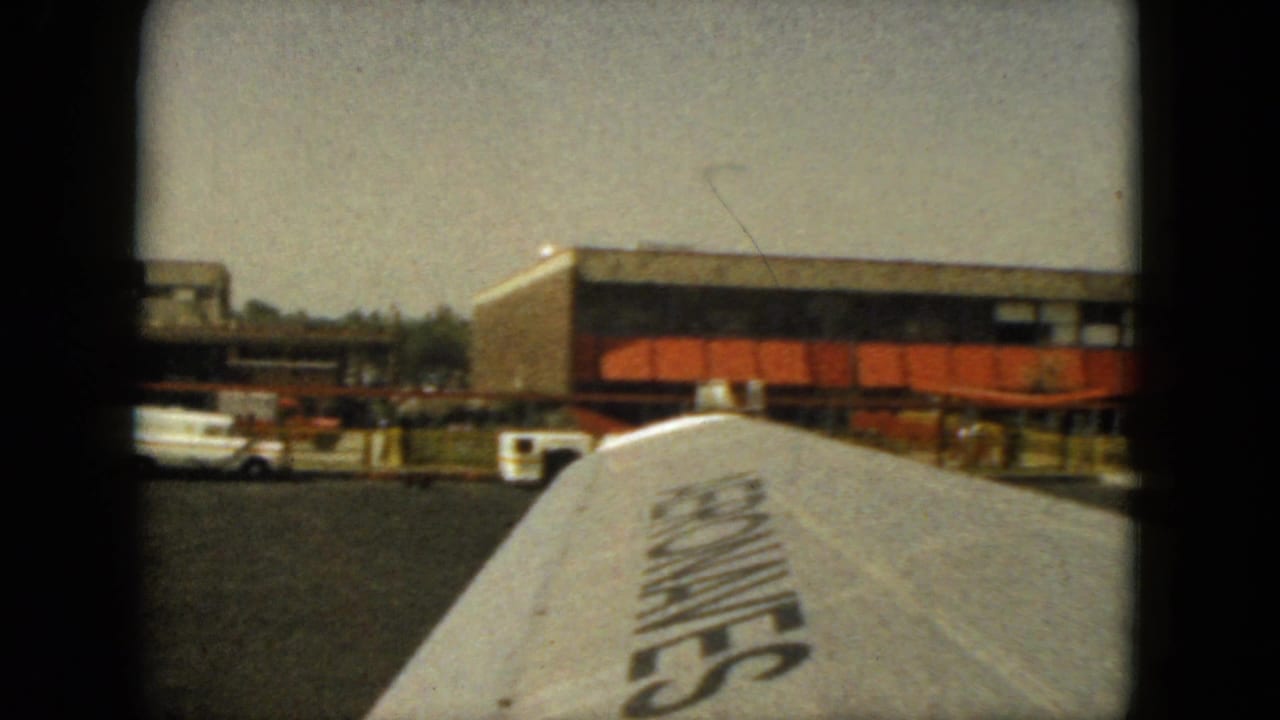Airplane wing in the foreground with terminal building, tarmac, and service vehicles beyond it.