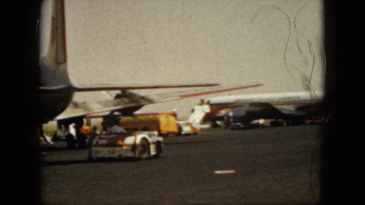 Commercial jet on an airport tarmac with ground vehicles and window framing in front.