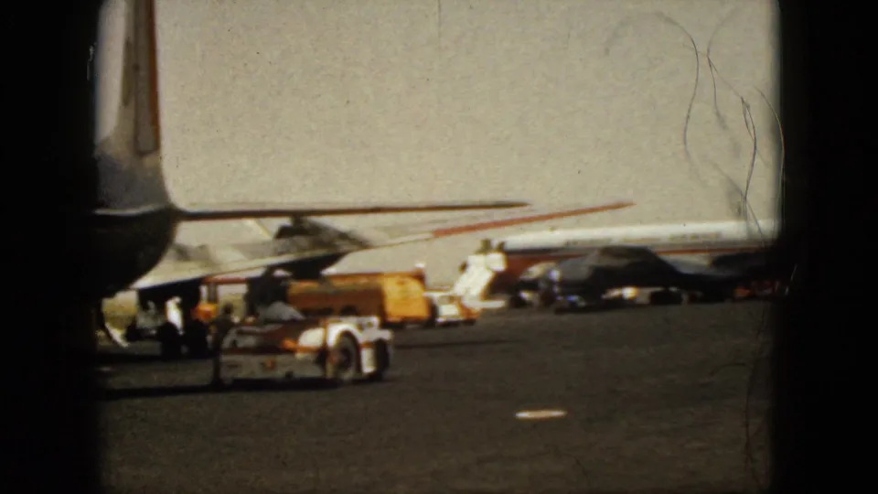 1962 Acapulco home-movie frame showing a parked commercial jet on the tarmac with service vehicles and dark terminal window framing.