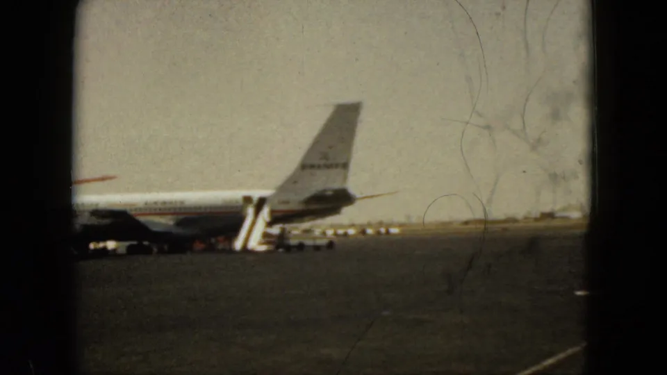 1962 Acapulco home-movie frame showing an airliner tail, mobile stairs, and airport apron viewed through dark window framing.