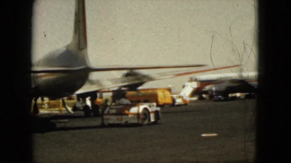 Brief 1962 Acapulco home-movie frame showing an aircraft tail on a tarmac with ground equipment and small people nearby.
