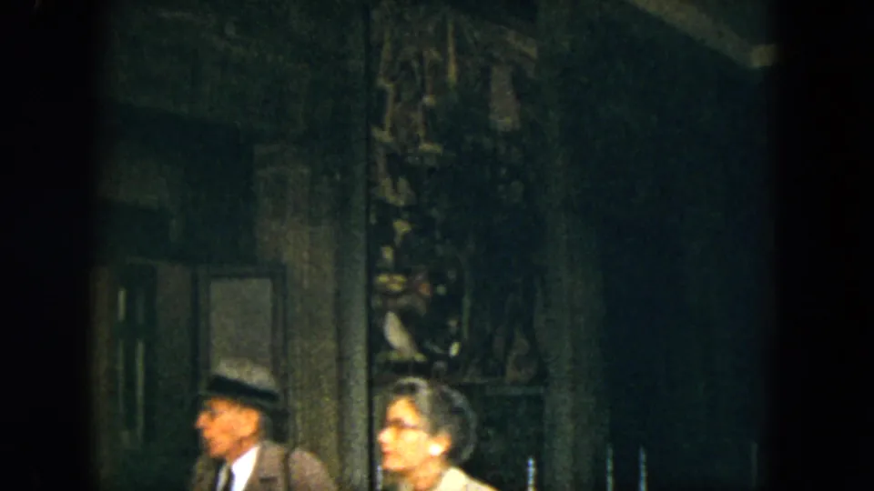 Adult visitors in 1960s dress move through a dark Acapulco interior captured on soft, grainy 8mm film.