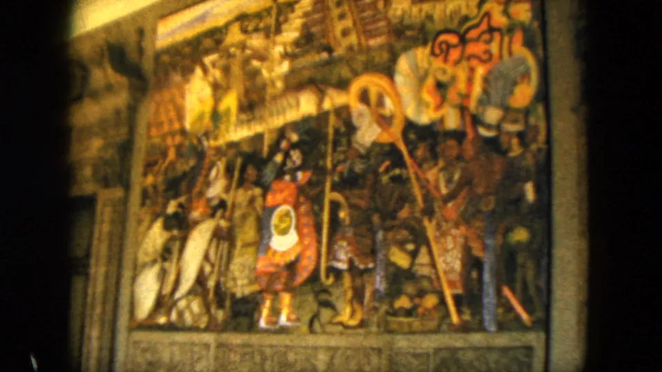 1962 Acapulco home-movie frame showing a detailed cultural mural with clustered figures, staffs, shield-like forms, and warm indoor color.