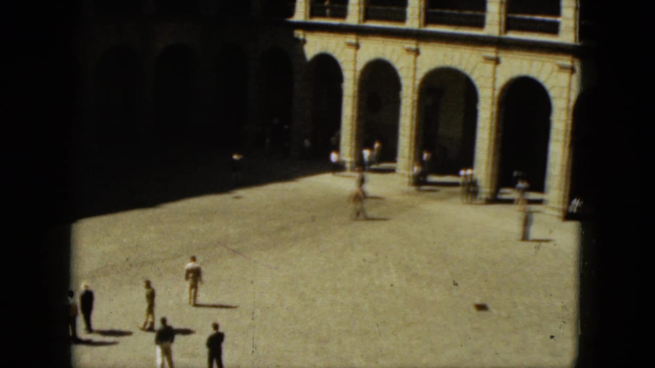 Pedestrians stand and walk across an open courtyard beside arched building fronts.