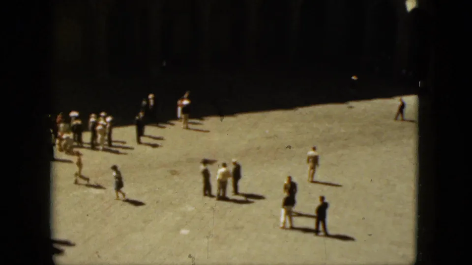 Wide 8mm frame of a busier Acapulco plaza, with pedestrians grouped across bright pavement near shaded colonnades.