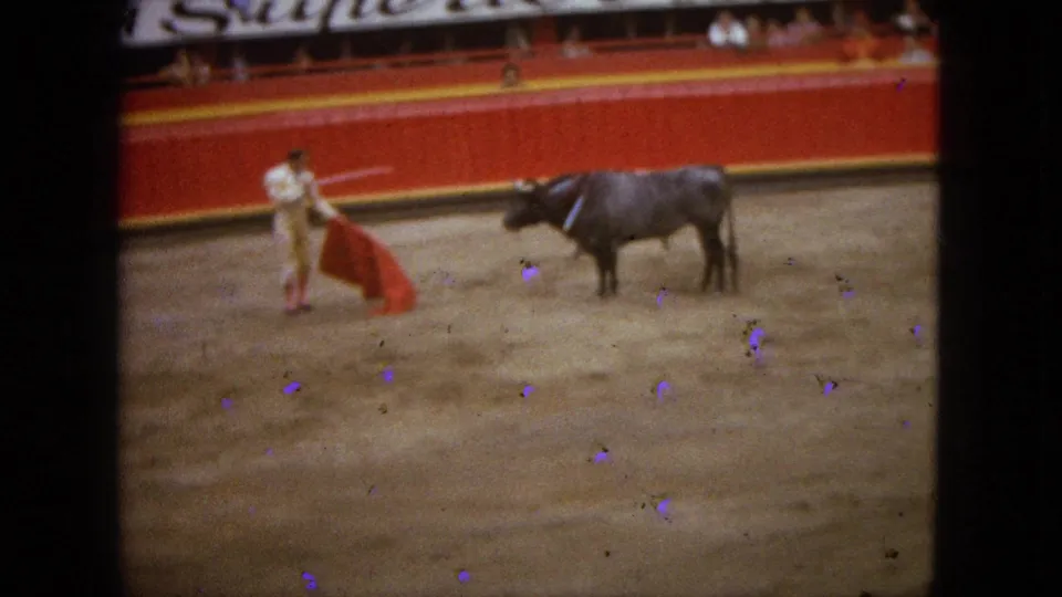 The matador holds the red cape low while the dark bull watches from the sandy ring in 1960s Acapulco footage.