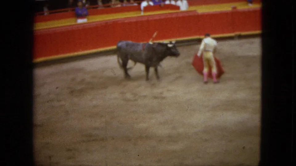 1962 Acapulco bullfighting frame with the matador holding a red cape near a dark bull beside red arena barriers.
