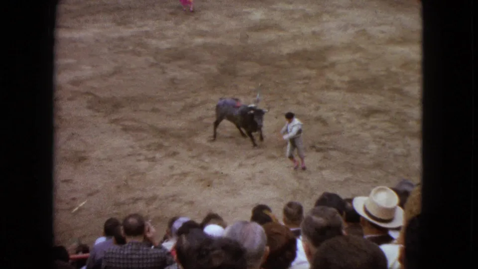 High-angle 1962 Acapulco bullfight frame showing the matador near the bull while audience members watch from the front rows.