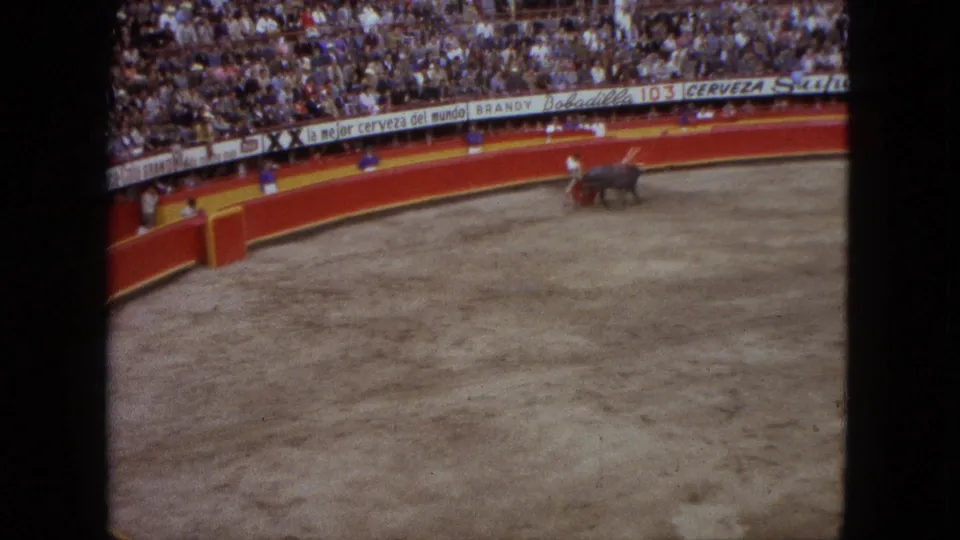 High-angle 1962 Acapulco bullring frame showing the bull and matador near the barrier with grandstand spectators and advertising boards.