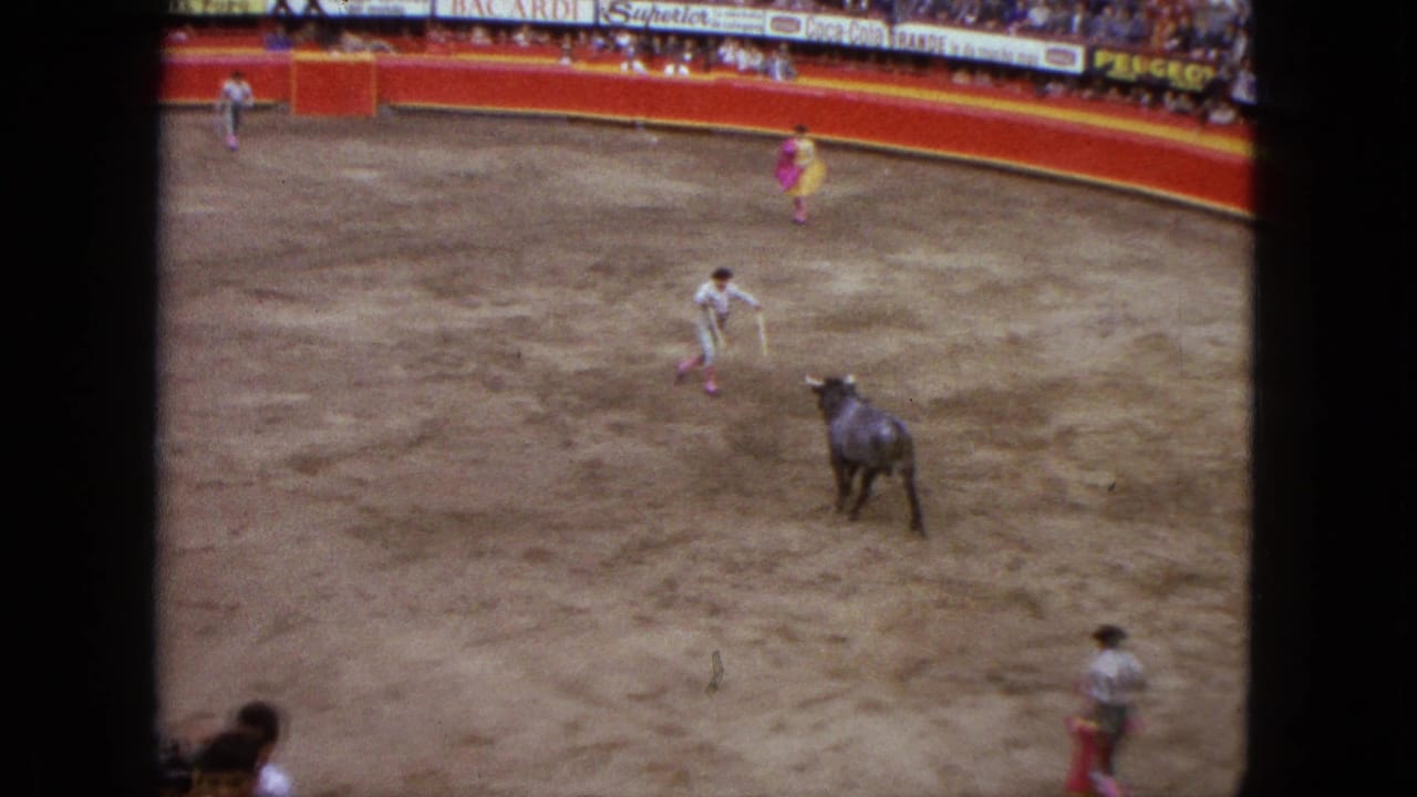 Elevated 1962 Acapulco arena view showing a bull approaching a matador, with cape holders spaced around the ring and spectators behind signage.