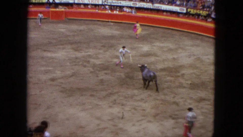Elevated 1962 Acapulco arena view showing a bull approaching a matador, with cape holders spaced around the ring and spectators behind signage.
