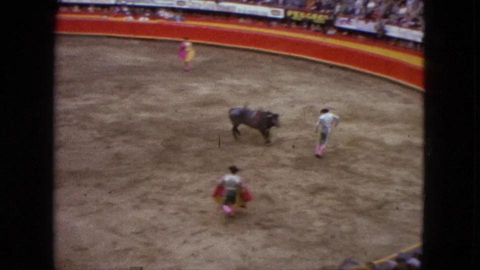 1962 Acapulco bullfighting scene with the bull moving across the ring, a matador nearby, and another cape holder kneeling in the foreground.