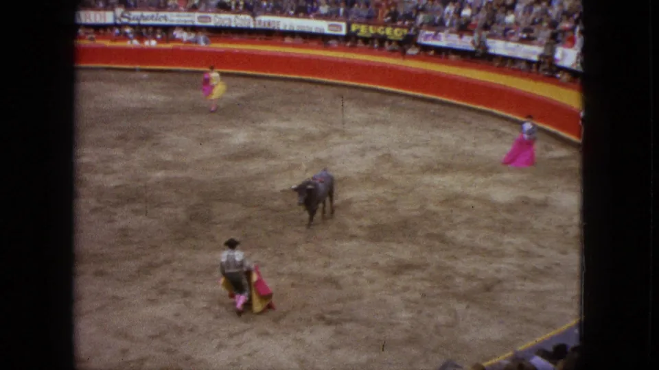 Wide 1962 Acapulco home-movie frame of a bull in the arena, matadors with pink and yellow capes, and spectators lining the stands.
