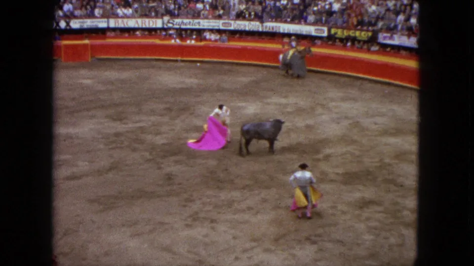 Wide 1962 Acapulco arena frame showing a matador with a pink cape, a black bull, and spectators behind the red ring barrier.