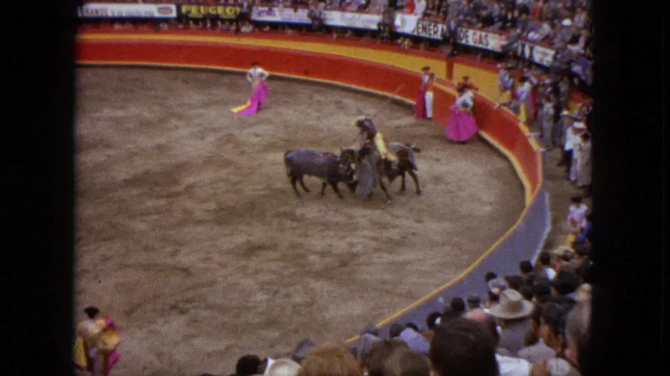 1962 Acapulco bullring scene with a black bull near mounted arena staff, matadors with capes, spectators, and visible ring advertising.