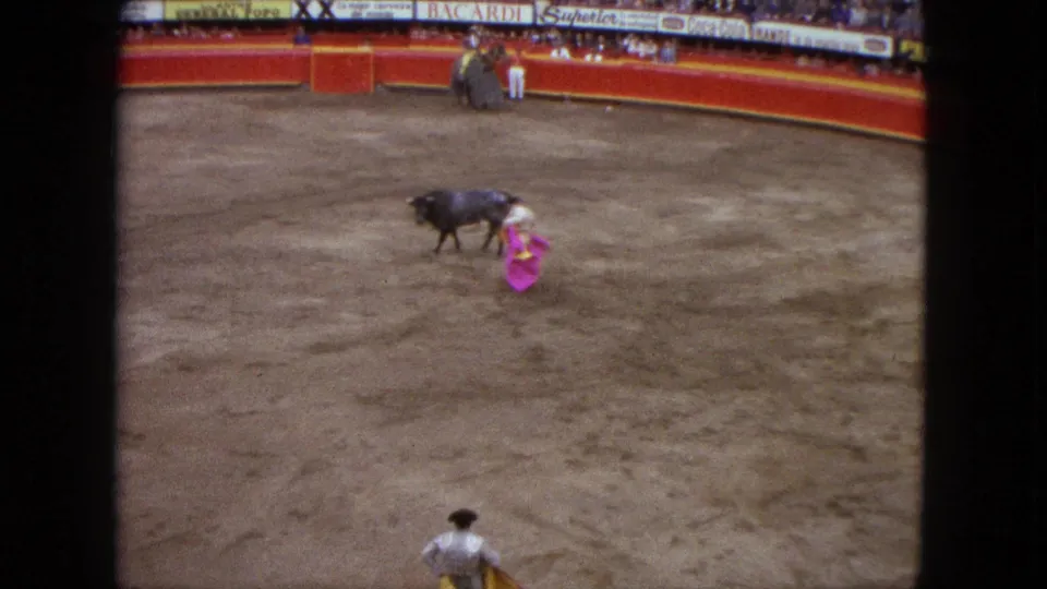 1962 Acapulco home-movie frame of a dark bull moving past a matador with a pink cape in a sandy arena.