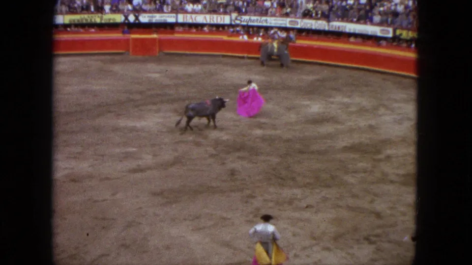 Wide 1962 Acapulco bullring view with a dark bull facing a matador in white holding a bright pink cape, seen from the stands.