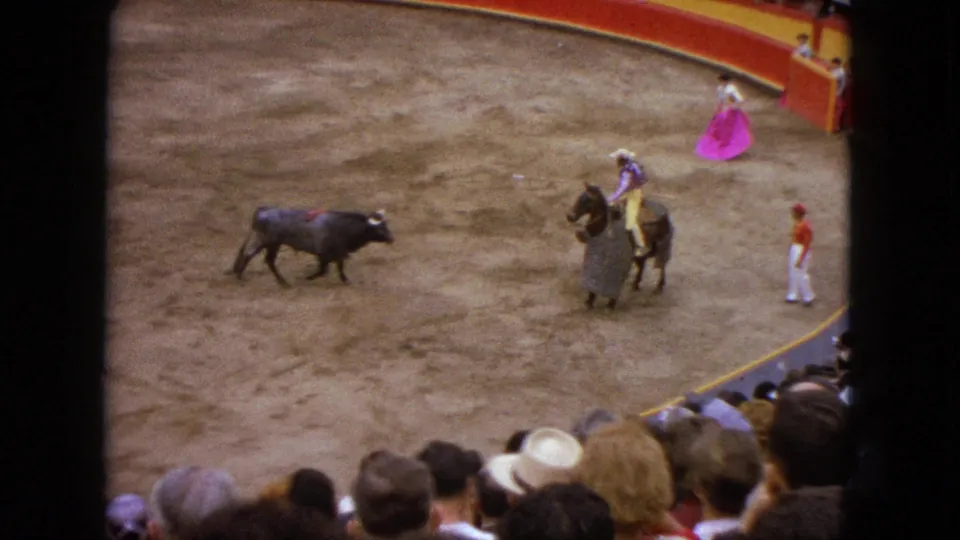 1962 Acapulco bullfighting arena scene with a dark bull, horseback rider, pink-caped matador, attendants, and spectators in the foreground.