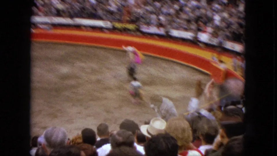 Crowded Acapulco bullring in 1962, with the matador and bull in motion and spectator heads visible in the foreground.