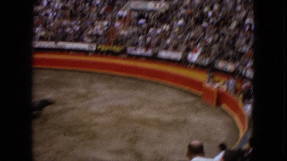 Handheld 8mm view across the Acapulco bullring, with a blurred animal at the edge, red barrier, and dense spectator stands.