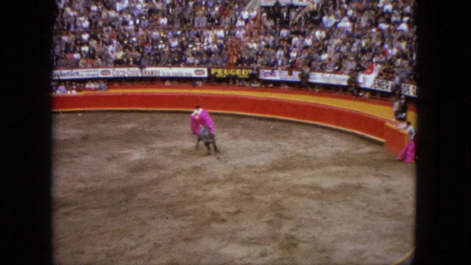 In the 1962 Acapulco bullring, a matador with a pink cape faces a bull while spectators fill the stands behind advertising boards.