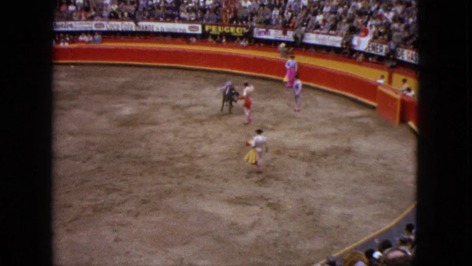 Elevated 8mm view of a matador engaging the bull with a red cape in the Acapulco arena, with another performer and crowd visible.
