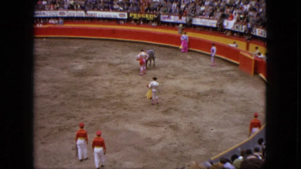 The bull remains in the sandy Acapulco ring with matadors nearby and attendants in red caps at the lower edge of the home-movie frame.