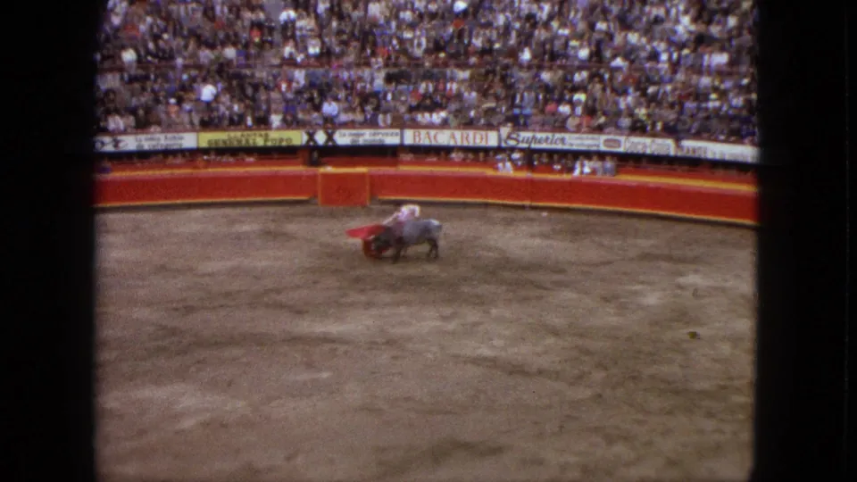 In the Acapulco bullring, the grey bull moves close to the matador's red cape as the packed stands and barrier signage remain visible.
