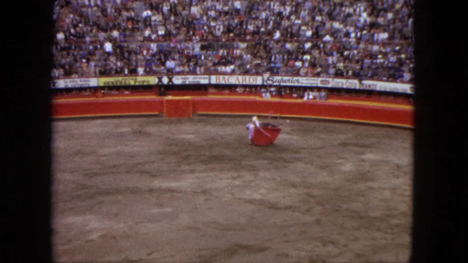 A wide spectator view from the 1962 Acapulco bullring shows the matador with red cape, the grey bull, packed seating, and sponsor boards.