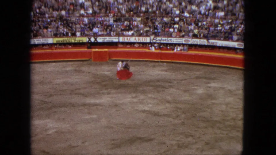 1962 Acapulco 8mm frame showing a matador in the ring as a bull approaches before packed stands and advertising boards.