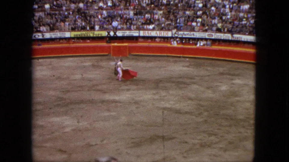 In 1962 Acapulco home-movie footage, a matador in light clothing presents a red cape to a bull in a dusty arena.