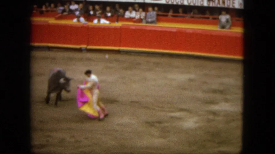 In 1962 Acapulco home-movie footage, a matador in white faces a small bull with a pink-and-yellow cape before seated spectators.