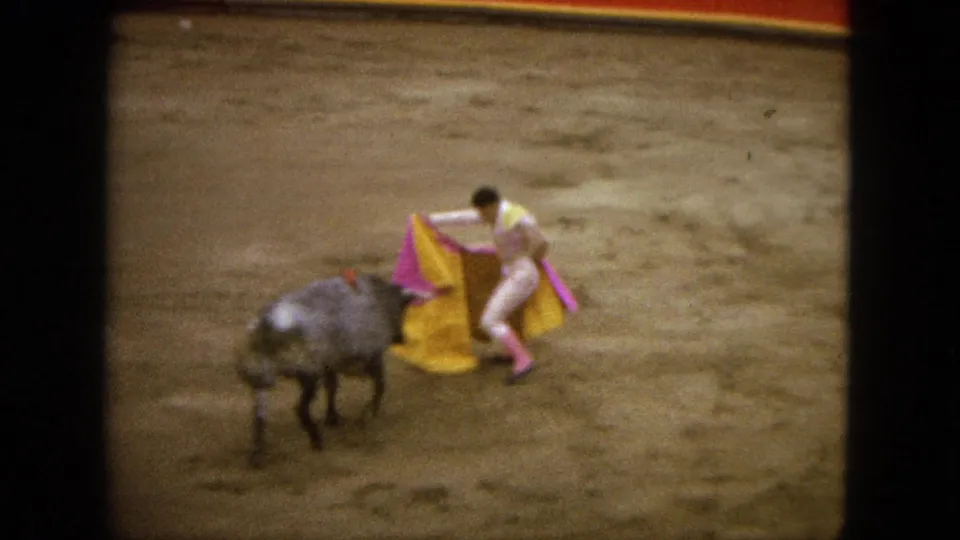 In 1962 Acapulco home-movie footage, a matador holds a pink and yellow cape near a grey bull inside a bullring.