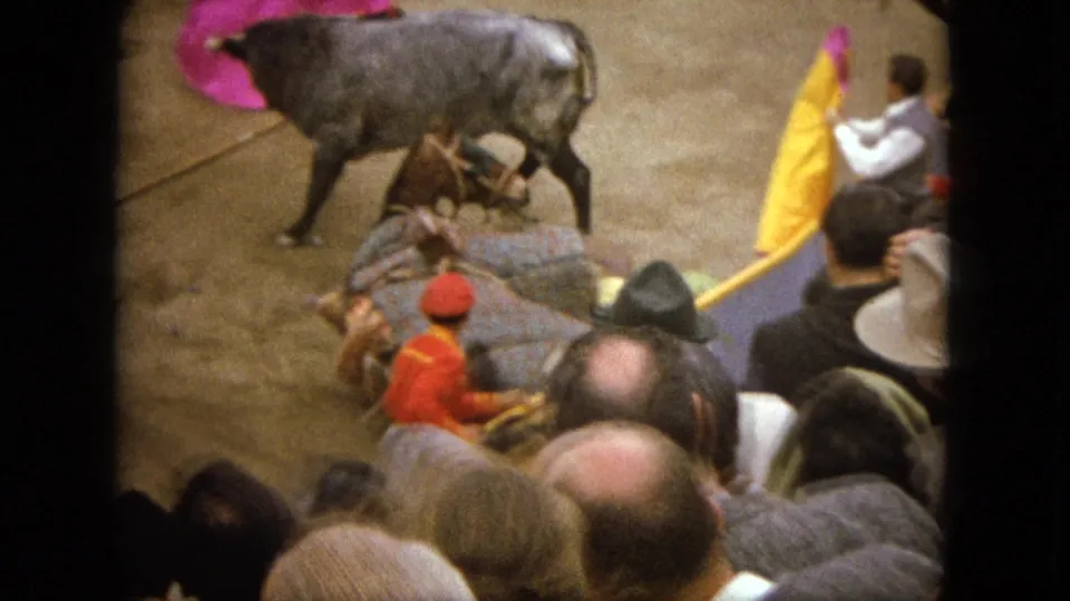 1962 Acapulco home-movie frame showing spectators in the stands as a grey bull moves near a matador's pink and yellow cape.