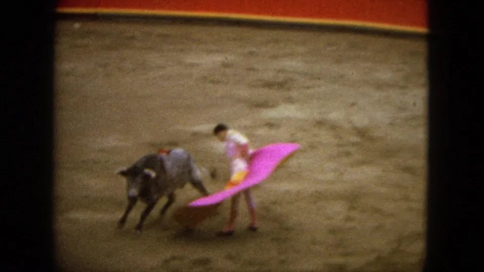 1962 Acapulco home-movie frame of a matador sweeping a pink cape while a grey bull turns on the sandy arena floor.