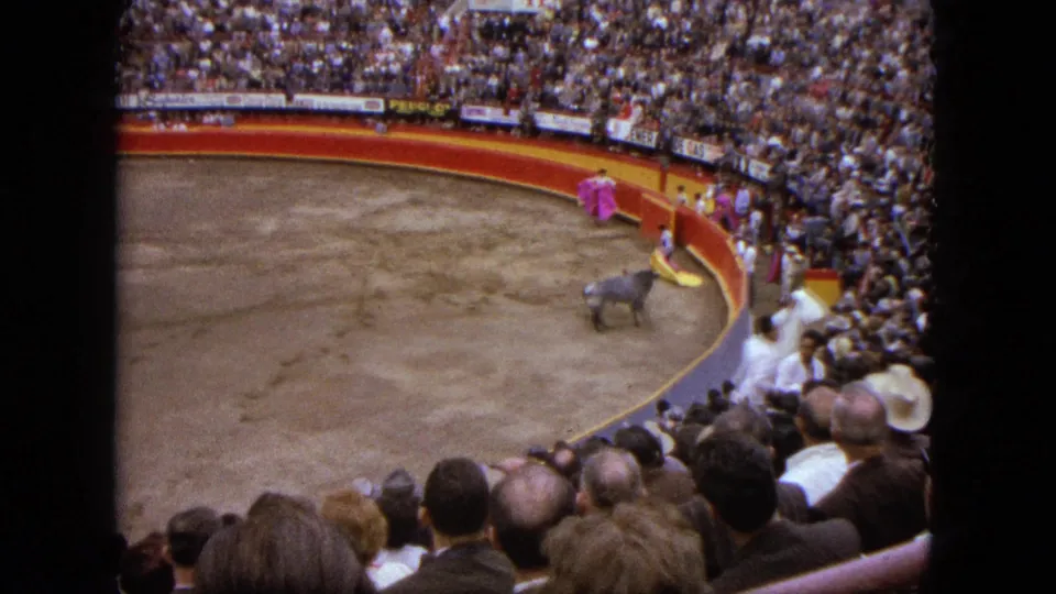 1962 Acapulco home-movie view from the stands showing a bull, matador, red arena barrier, and packed spectators.