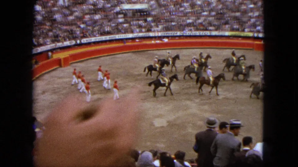 Home-movie frame from the Acapulco arena with a foreground hand, riders, performers, and spectators in view.
