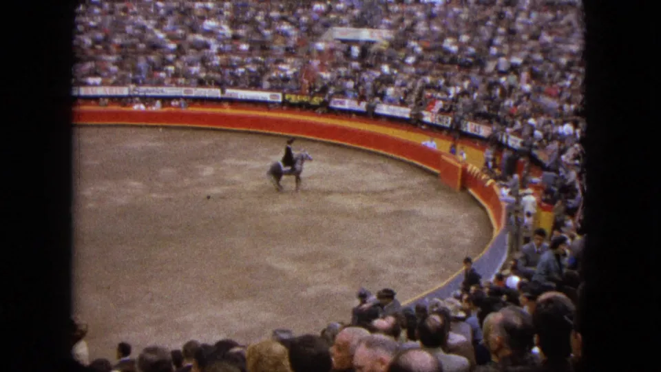 A rider on a grey horse crosses the sandy ring in Acapulco, with dense 1960s spectator seating behind.