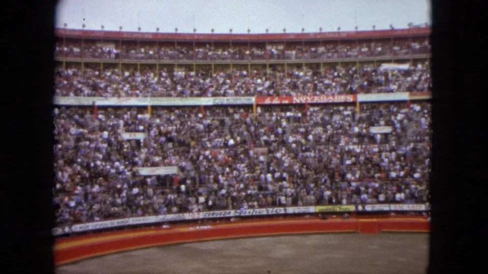 1962 Acapulco home-movie frame showing densely filled stadium seating around a curved arena wall with visible advertising banners.