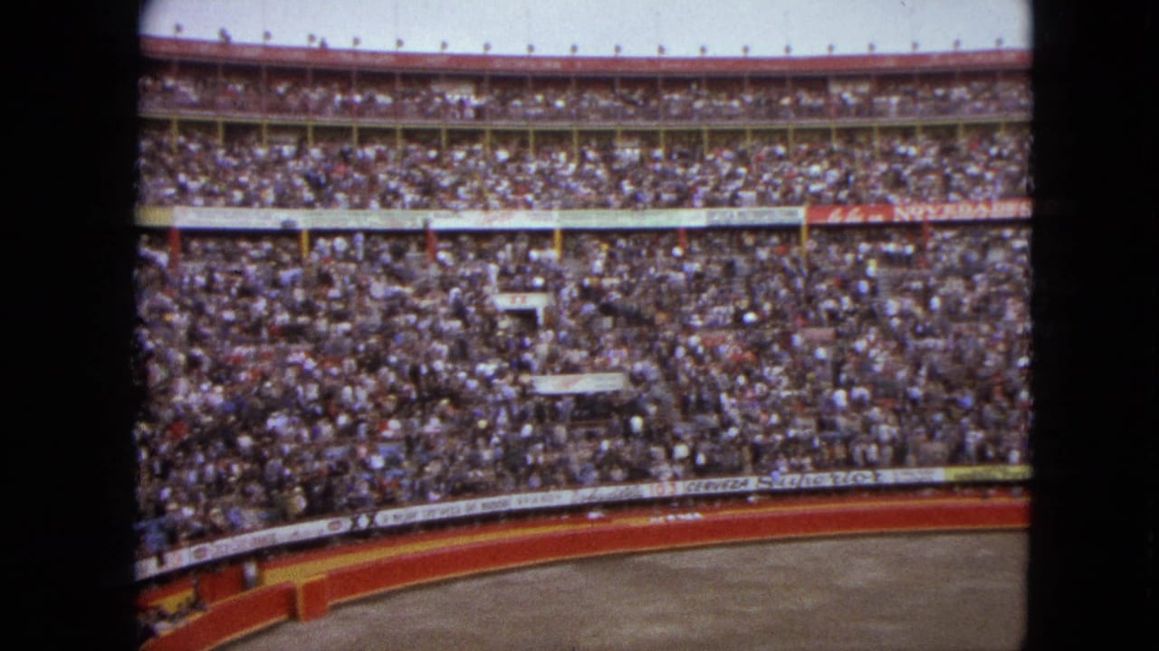 Wide 8mm view of a packed Acapulco stadium crowd in 1962, with red arena walls and advertising along the lower railing.
