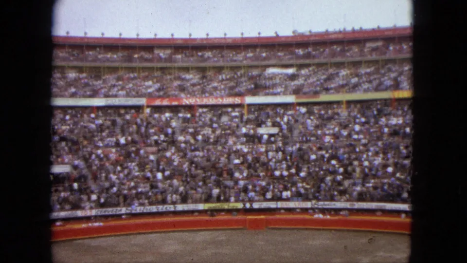 The home-movie camera looks across a packed Acapulco arena toward rows of spectators and the red barrier below.
