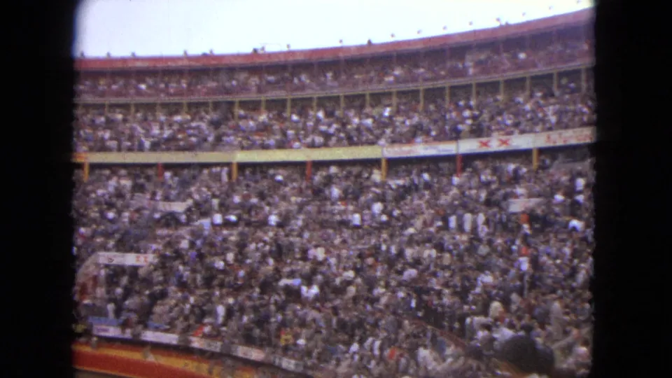 Handheld 8mm view across the Acapulco bullring stands, showing a large crowd and partial arena barrier in the foreground.