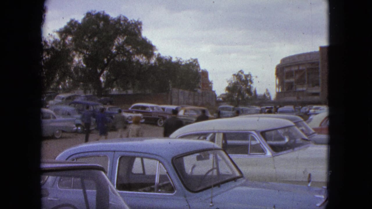 Classic cars parked near trees and a rounded arena building in the background.
