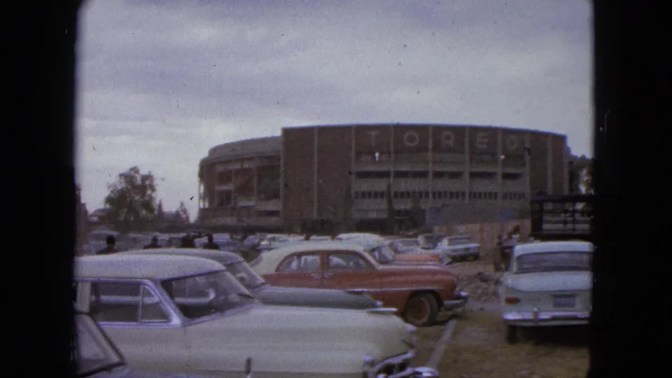 1962 Acapulco home-movie view of the TOREO-marked building with classic cars, a red sedan, and people in the lot.