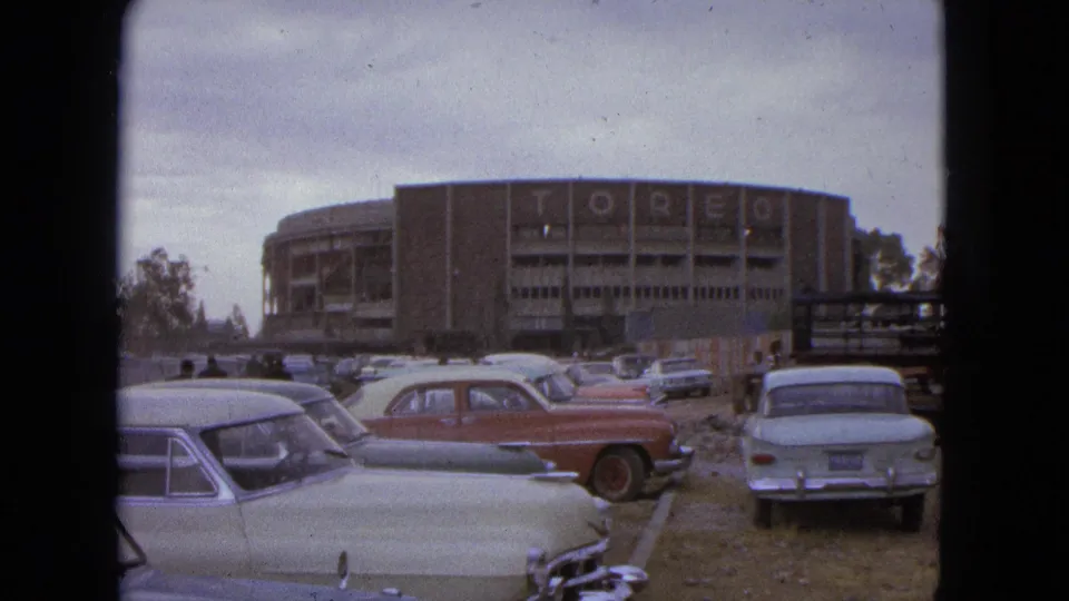 1962 Acapulco frame showing a red sedan, pale parked cars, pedestrians, and a large building with TOREO lettering.
